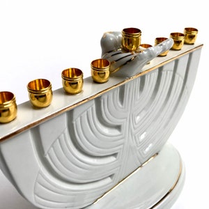 May include: White ceramic menorah with gold accents and a stylized tree design. The menorah has nine candle holders, each with a gold rim. The menorah is decorated with a gold-painted bird and a hand.