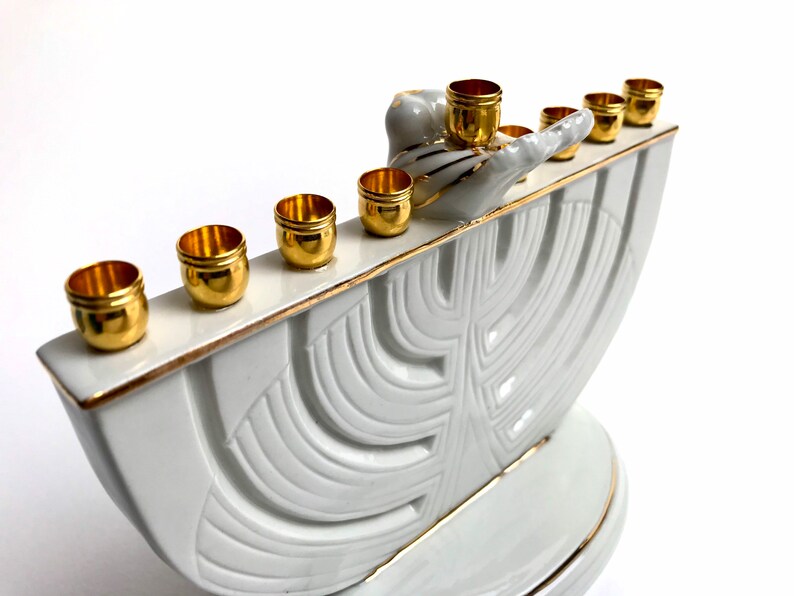 May include: White ceramic menorah with gold accents and a stylized tree design. The menorah has nine candle holders, each with a gold rim. The menorah is decorated with a gold-painted bird and a hand.