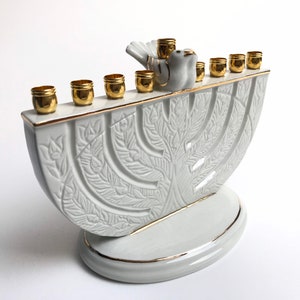 May include: White ceramic menorah with a gold trim and a dove design. The menorah has nine candle holders and a base with a tree design.
