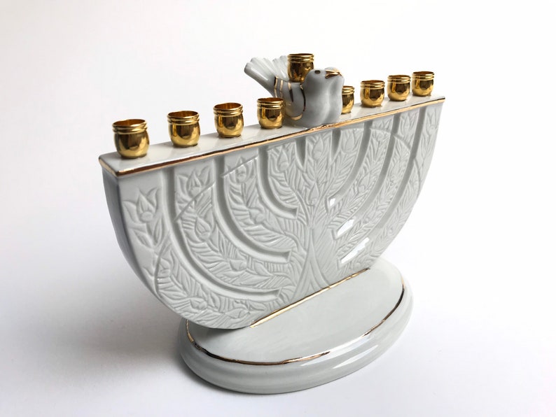 May include: White ceramic menorah with a gold trim and a dove design. The menorah has nine candle holders and a base with a tree design.