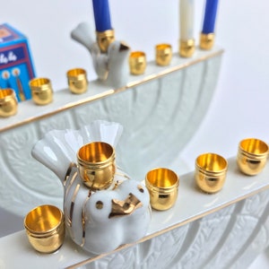May include: A white porcelain menorah with gold accents and dove designs. The menorah has nine candle holders and is decorated with a floral pattern. The menorah is a traditional Jewish symbol of the Festival of Lights.