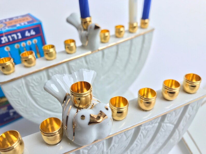 May include: A white porcelain menorah with gold accents and dove designs. The menorah has nine candle holders and is decorated with a floral pattern. The menorah is a traditional Jewish symbol of the Festival of Lights.
