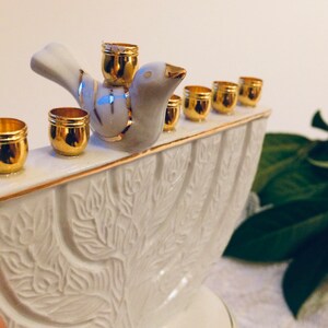 May include: White ceramic menorah with a gold trim and a dove figurine on top. The menorah has nine candle holders, each with a gold rim.