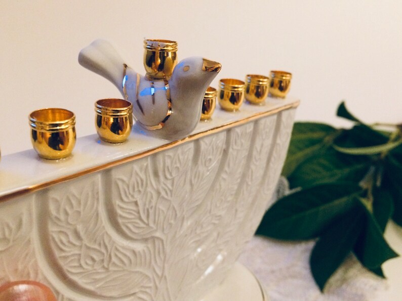 May include: White ceramic menorah with a gold trim and a dove figurine on top. The menorah has nine candle holders, each with a gold rim.
