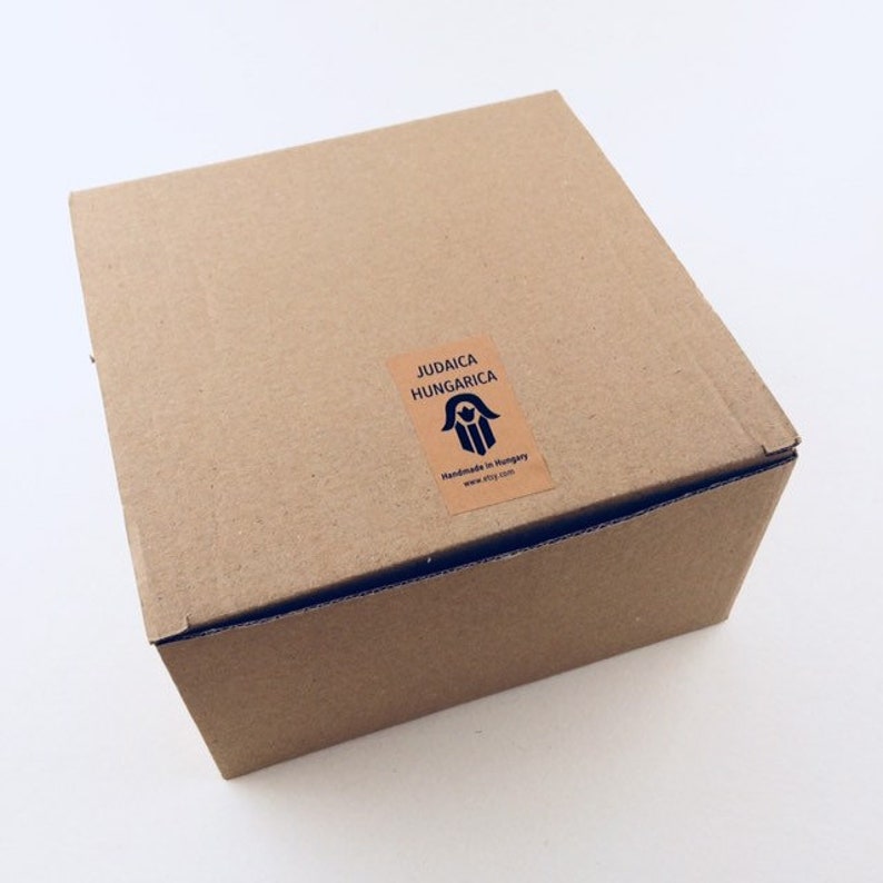 May include: A brown cardboard box with a label that reads "Judaica Hungarica" and features a hamsa hand symbol. The label also states "Handmade in Hungary" and includes a website address.
