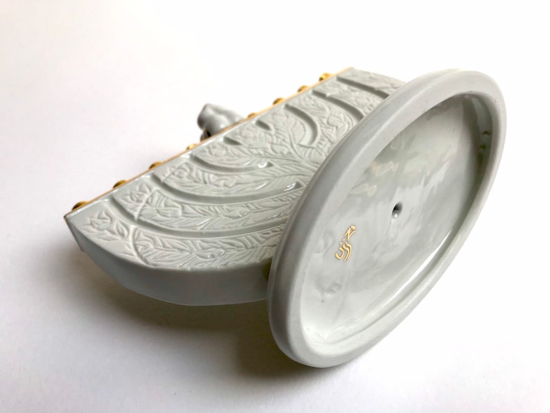 May include: White ceramic menorah with gold accents and a floral design. The base is oval shaped and has a small hole in the center. The menorah is marked with the word "כשר" in Hebrew.