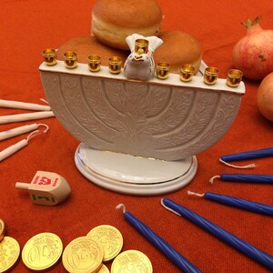 May include: A white ceramic menorah with gold accents and a dove figurine on top. The menorah is lit with eight golden candles and surrounded by gelt, sufganiyot, and pomegranates.