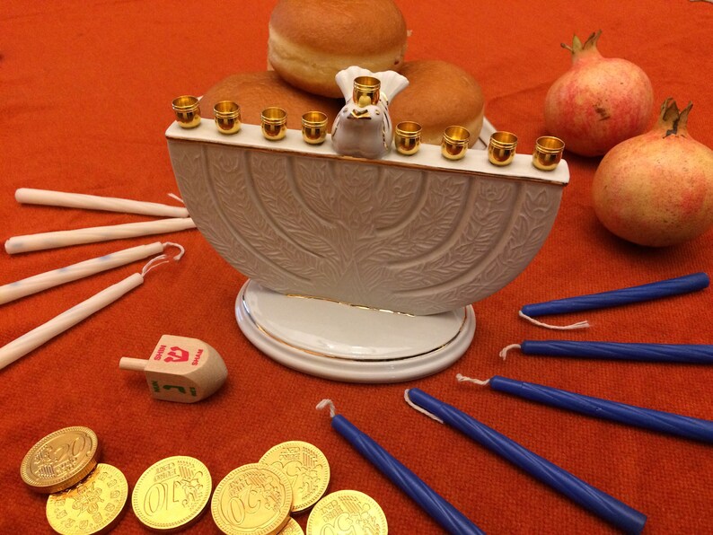 May include: A white ceramic menorah with gold accents and a dove figurine on top. The menorah is lit with eight golden candles and surrounded by gelt, sufganiyot, and pomegranates.