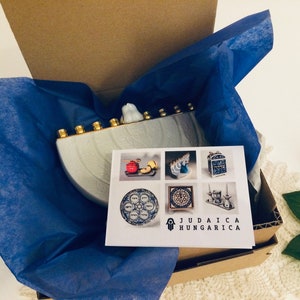 May include: A white ceramic menorah with gold accents, packaged in a brown box with blue tissue paper. The menorah is shaped like a half-circle and has seven candle holders. The box has a card with the text "Judaica Hungarica" and images of other menorahs and Judaica items.