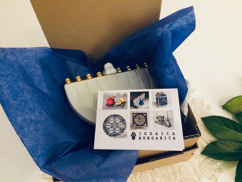May include: A white ceramic menorah with gold accents, packaged in a brown box with blue tissue paper. The menorah is shaped like a half-circle and has seven candle holders. The box has a card with the text "Judaica Hungarica" and images of other menorahs and Judaica items.