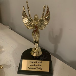 Peut inclure: Un trophée doré avec une figure ailée tenant une torche. Le socle est en marbre noir avec une plaque dorée sur laquelle est inscrit "High School Graduation Class of 2023".