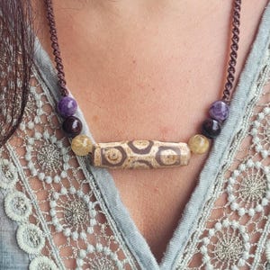 Può includere: Una collana con cordino marrone con un ciondolo cilindrico con un motivo geometrico. La collana include perline di pietre preziose viola, marroni e gialle. La modella indossa un top azzurro con un design in pizzo.