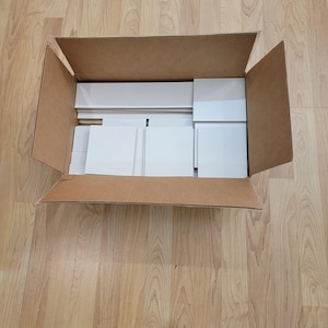 May include: An open cardboard box containing several white, rectangular pieces, possibly for construction or assembly. The box is on a wooden floor, and the pieces are neatly arranged inside. The box is brown.