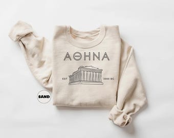 Peut inclure: Un sweat-shirt beige avec un motif graphique noir du Parthénon à Athènes, en Grèce. Le texte "AOHNA EST 3000 BC" est imprimé sur le sweat-shirt.