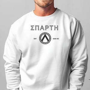 Könnte beinhalten: Weißer Pullover mit einem schwarzen Grafikdruck des Wortes "ΣΠAPTH" und einem Schild mit einem griechischen Schlüsselförmigen Design. Der Text "EST 3000 BC" ist unter dem Schild gedruckt.