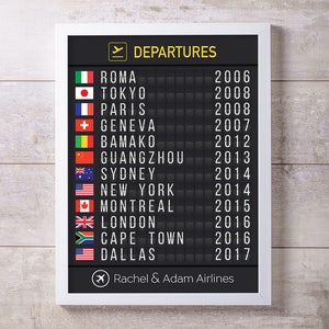 Puede incluir: Un tablero de salidas de aeropuerto en blanco y negro con un marco blanco. El tablero muestra una lista de ciudades y a&ntilde;os, incluyendo Roma, Tokio, Par&iacute;s, Ginebra, Bamako, Guangzhou, Sydney, Nueva York, Montreal, Londres, Ciudad del Cabo y Dallas. El tablero tambi&eacute;n incluye un peque&ntilde;o icono de avi&oacute;n y el texto "Rachel & Adam Airlines".