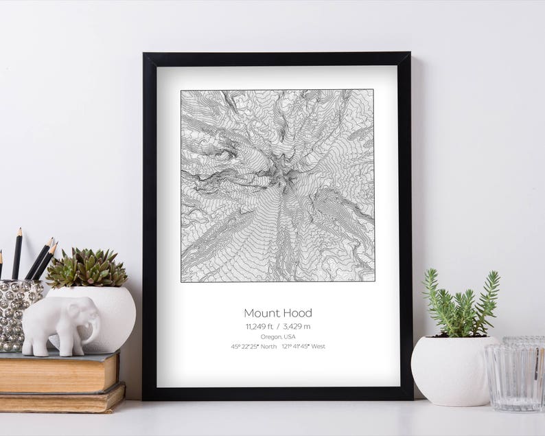 May include: A black framed print of a topographic map of Mount Hood, Oregon. The print includes the mountain's elevation, 11,249 ft / 3429 m, and coordinates, 45&deg; 22'25" North 121&deg; 45'47" West.