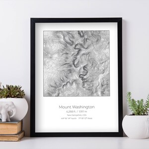 May include: A black and white topographic map print of Mount Washington in New Hampshire, USA. The print shows the elevation contours of the mountain, with the peak at 6,288 feet (1,917 meters). The print is framed in a black frame and has a white mat.