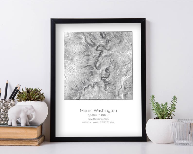 May include: A black and white topographic map print of Mount Washington in New Hampshire, USA. The print shows the elevation contours of the mountain, with the peak at 6,288 feet (1,917 meters). The print is framed in a black frame and has a white mat.