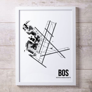 May include: A black and white print of the Boston Logan International Airport (BOS) layout. The print is framed in a white frame and has the text "BOS" and "BOSTON, MASSACHUSETTS" below the airport layout.