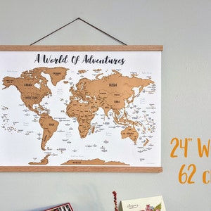 May include: A gold world map with the text "A World of Adventures" printed at the top. The map is framed with a light brown wooden frame and is hanging on a light gray wall. The map is 24 inches wide.