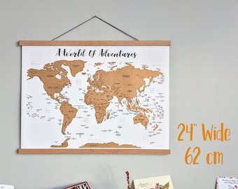 May include: A gold world map with the text "A World of Adventures" printed at the top. The map is framed with a light brown wooden frame and is hanging on a light gray wall. The map is 24 inches wide.