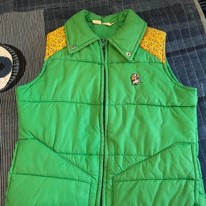 May include: A green puffer vest with a yellow and floral patterned fabric on the shoulders. The vest has a zipper closure and a small embroidered dog on the front.