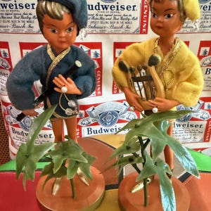 Op de afbeelding: Twee vintage beeldjes van kinderen, een in een blauwe outfit met een fluit en de andere in een gele outfit met een harp. Elk beeldje houdt een groene plant vast op een ronde, bruine basis. De achtergrond toont een Budweiser bieradvertentie.