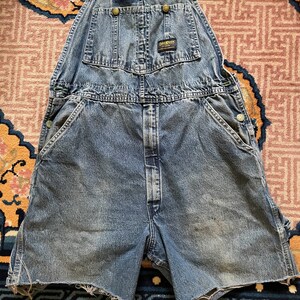 May include: A pair of blue denim dungarees with a faded look. The dungarees have a short leg length and are cut off at the thigh. The dungarees have a brand label that reads "Dickies" on the front.