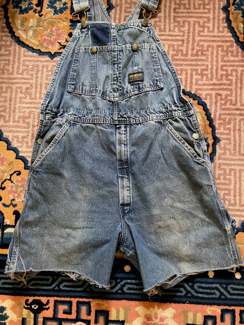 May include: A pair of blue denim dungarees with a faded look. The dungarees have a short leg length and are cut off at the thigh. The dungarees have a brand label that reads "Dickies" on the front.
