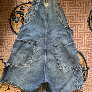 May include: A pair of blue denim dungarees with a faded look. The dungarees have a distressed hem and a small patch on the front with the text "Carhartt" in white.