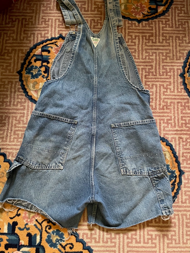 May include: A pair of blue denim dungarees with a faded look. The dungarees have a distressed hem and a small patch on the front with the text "Carhartt" in white.