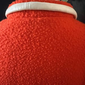 May include: Close-up of a bright orange coat with a textured surface. The coat has a white collar. The image is focused on the back of the coat, showing the collar and the textured fabric.