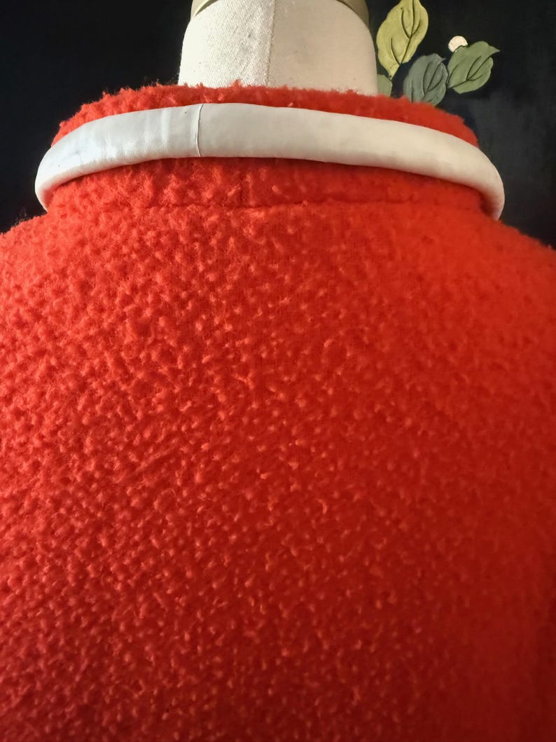 May include: Close-up of a bright orange coat with a textured surface. The coat has a white collar. The image is focused on the back of the coat, showing the collar and the textured fabric.