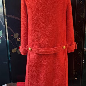 May include: A bright red, textured coat with a white collar and cuffs. The coat features a belt with two gold buttons and matching buttons on the sleeves. The coat is displayed on a mannequin.