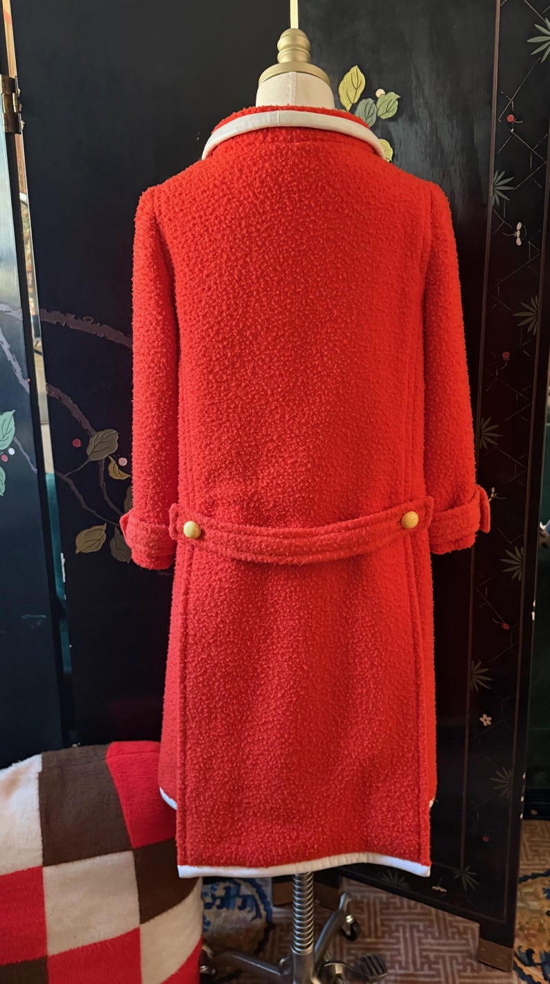 May include: A bright red, textured coat with a white collar and cuffs. The coat features a belt with two gold buttons and matching buttons on the sleeves. The coat is displayed on a mannequin.