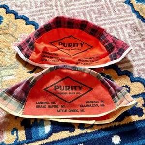 May include: Two orange welder caps with black plaid brims. Each cap features a black diamond logo with the text PURITY CYLINDER GASES INC. and a list of locations including Lansing, Grand Rapids, Battle Creek, Warsaw, Kalamazoo, and Mt. Pleasant.