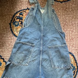 May include: A pair of light blue denim dungarees with a frayed hem. The dungarees have two large pockets on the back and a small pocket on the front. The dungarees are made of a soft, comfortable denim.