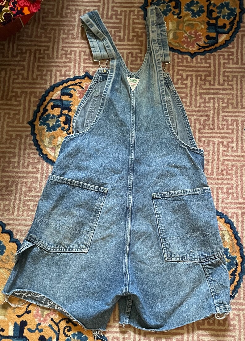 May include: A pair of light blue denim dungarees with a frayed hem. The dungarees have two large pockets on the back and a small pocket on the front. The dungarees are made of a soft, comfortable denim.