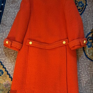 May include: A vintage orange coat with a textured fabric. The coat features a white collar and cuffs, a matching belt detail with two yellow buttons, and yellow buttons on the sleeves. The coat is a knee-length style.