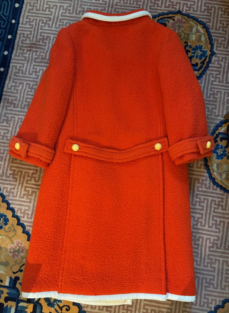 May include: A vintage orange coat with a textured fabric. The coat features a white collar and cuffs, a matching belt detail with two yellow buttons, and yellow buttons on the sleeves. The coat is a knee-length style.