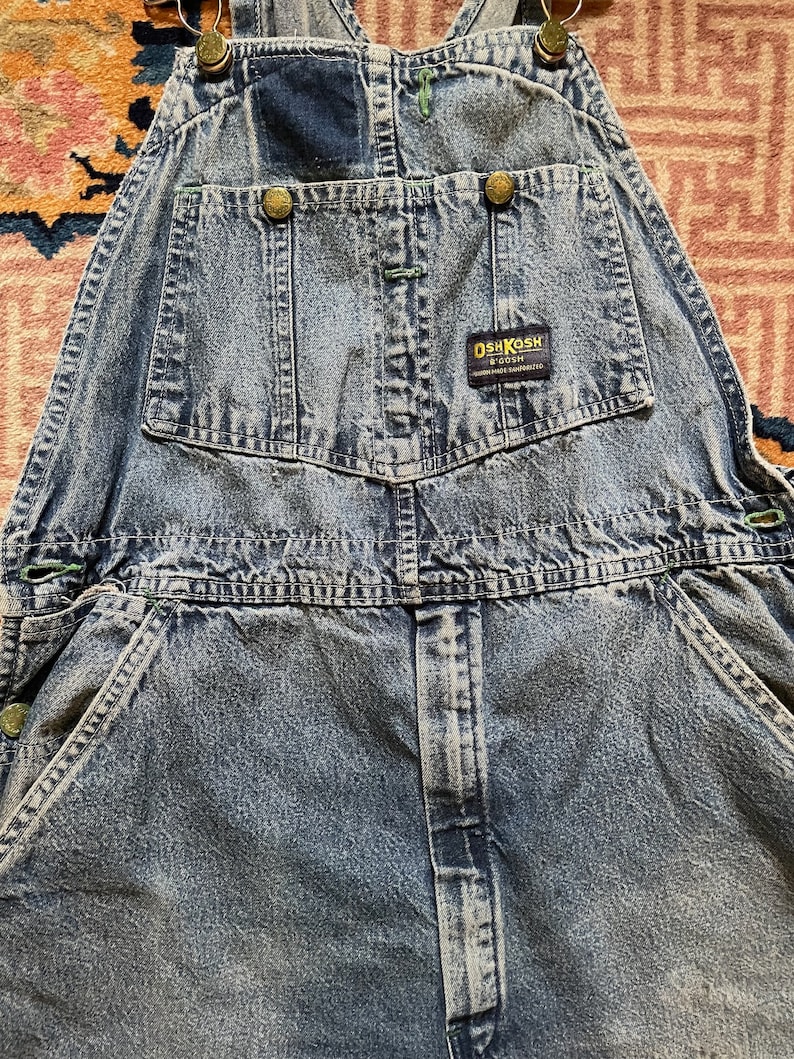 May include: Light blue denim dungarees with a green and blue patch on the front pocket. The dungarees have a brown leather label with the text "OSHKOSH B'GOSH" and "MADE IN THE USA".
