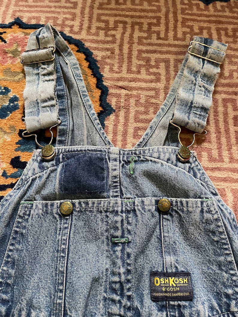 May include: Light blue denim dungarees with adjustable straps and a patch on the front that reads "OshKosh B'Gosh Union Made Sanforized".
