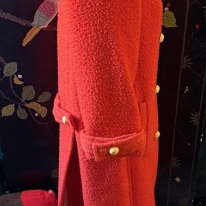 May include: A bright red, textured coat with a white collar and gold buttons. The coat has a double-breasted design and a tailored fit. The sleeves are cuffed, and the coat is knee-length.