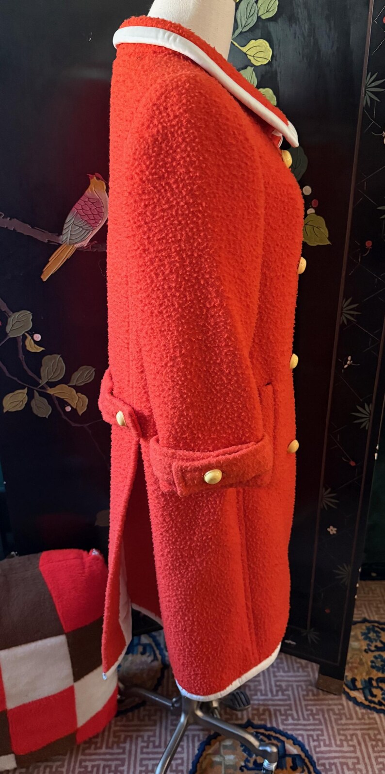 May include: A bright red, textured coat with a white collar and gold buttons. The coat has a double-breasted design and a tailored fit. The sleeves are cuffed, and the coat is knee-length.