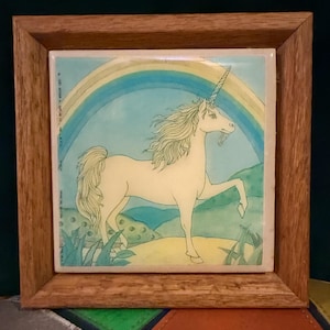 Puede incluir: Azulejo enmarcado con un unicornio y un arcoíris. El unicornio blanco tiene una melena y cola fluidas, sobre un paisaje verde. El marco es de madera.