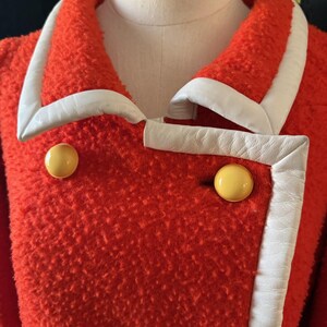 May include: A vintage red coat with white trim and large yellow buttons. The coat has a textured fabric and a double-breasted design. The white trim outlines the collar and front edges, creating a striking contrast.