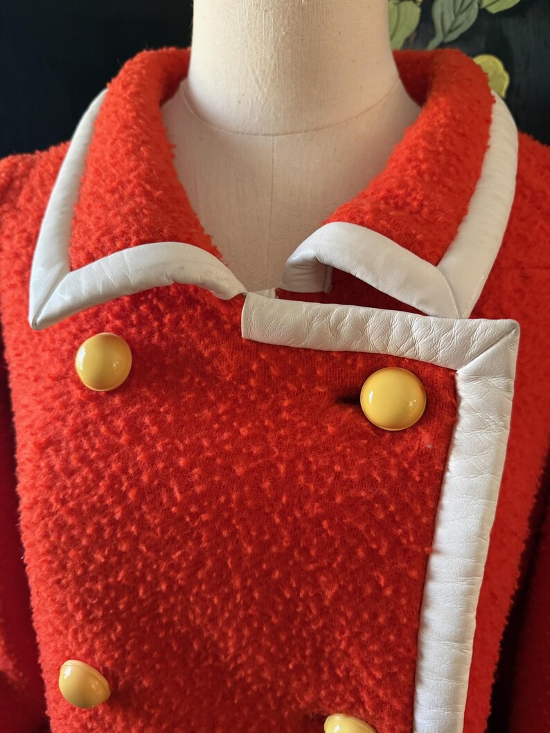 May include: A vintage red coat with white trim and large yellow buttons. The coat has a textured fabric and a double-breasted design. The white trim outlines the collar and front edges, creating a striking contrast.