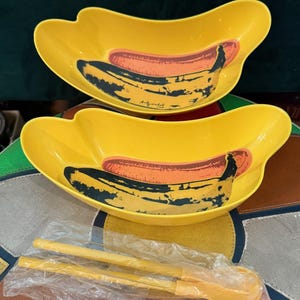 May include: Two yellow, banana-shaped serving dishes with a hot dog and banana design. The dishes are paired with yellow serving utensils in a plastic bag. The design is a pop art style.