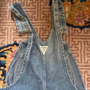 May include: A pair of light blue denim dungarees with adjustable straps. The dungarees have a brand label that reads "OshKosh B'Gosh" and "Vest-Tex".